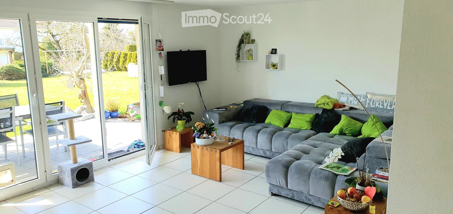 Bright living room with large sliding glass doors opening to a garden, grey sectional sofa with green cushions, wooden coffee table, cat tree, cat house, small table with flower pot, and a television mounted on the wall.