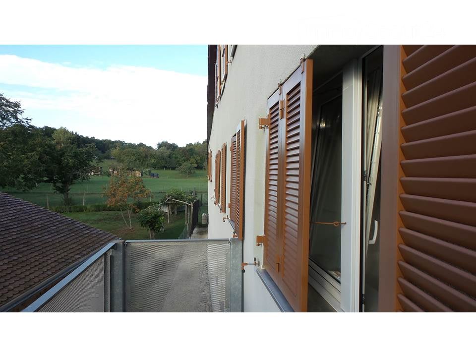Balcony with wooden shutters, overlooking a green field and trees under a partly cloudy sky.