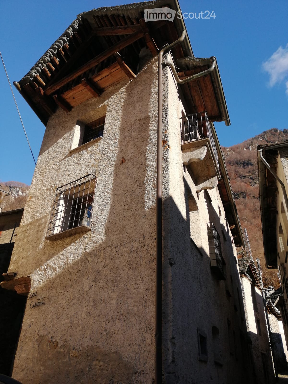 A rustic stone building with wooden balconies and metal railings, set against a clear blue sky and mountainous backdrop.