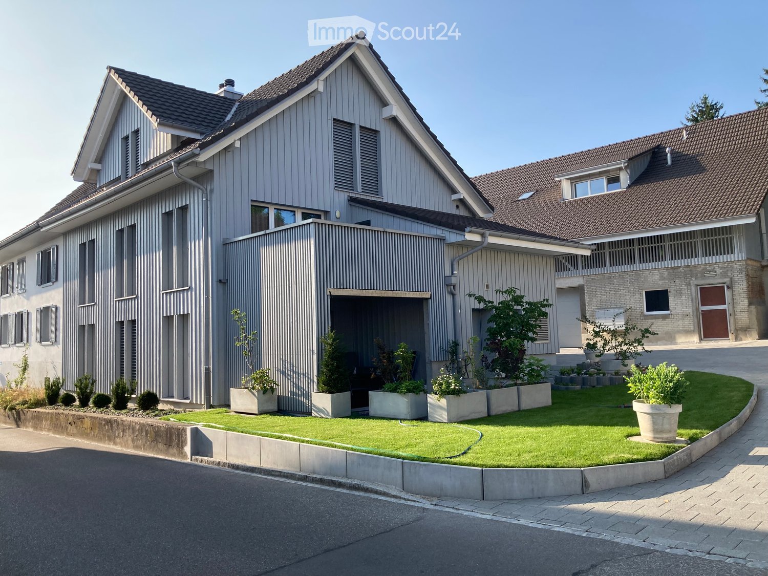 Modern two-story house with grey siding, multiple windows, and a garage entrance. The front yard features a well-maintained lawn, potted plants, and a small garden area.
