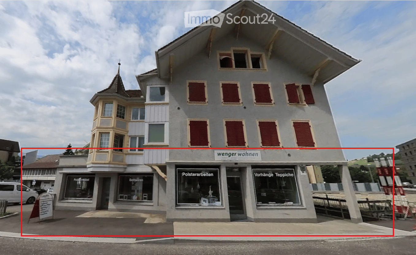 3 story building, gray exterior, red shutters, storefronts on the ground floor, sign 'wenger wohnen', sign 'Polsterarbeiten', sign 'Vorhänge Teppiche'
