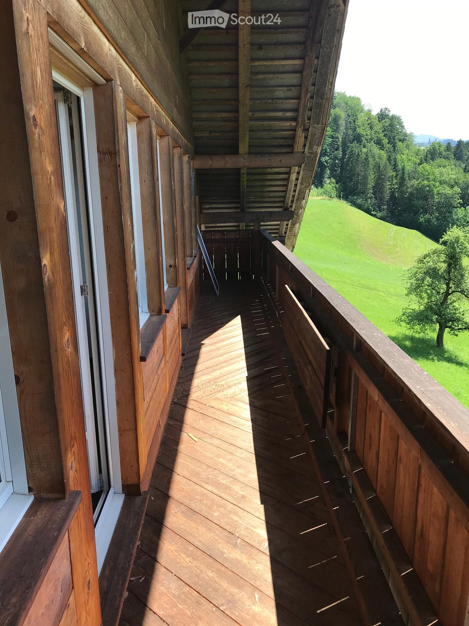 Wooden balcony with railing, overlooking a green field and trees.