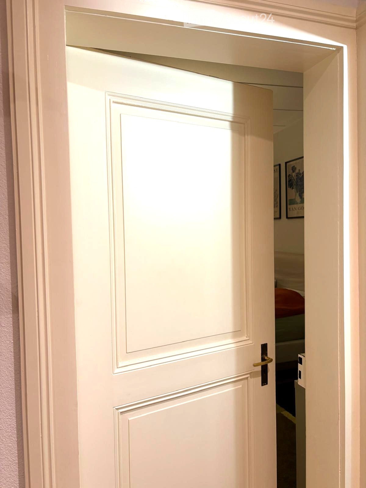 Open doorway leading to another room, with visible interior elements like a bed and artwork in the background.