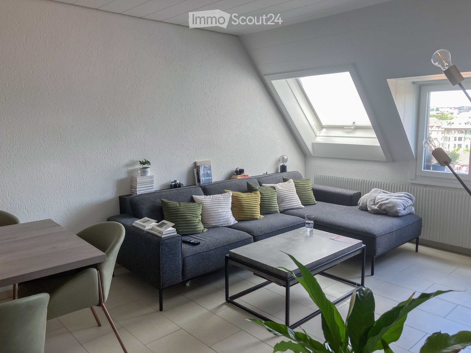 Modern living room with grey sectional sofa, coffee table, dining table, chairs, books, and decorative pillows.