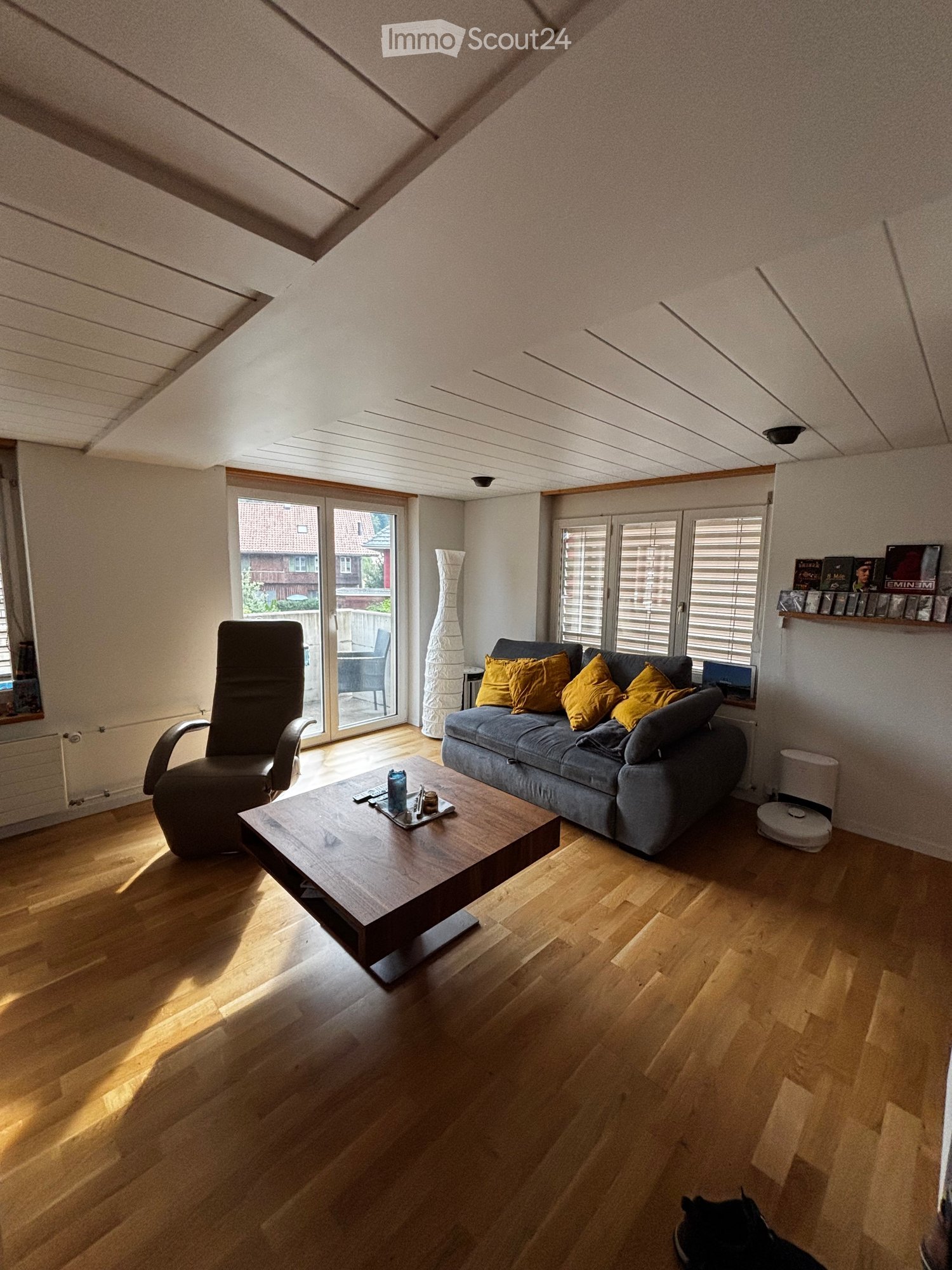 Bright living room with wooden flooring, large windows, and a sliding glass door leading to a patio. Features a grey sofa with yellow cushions, a wooden coffee table, and a black armchair.