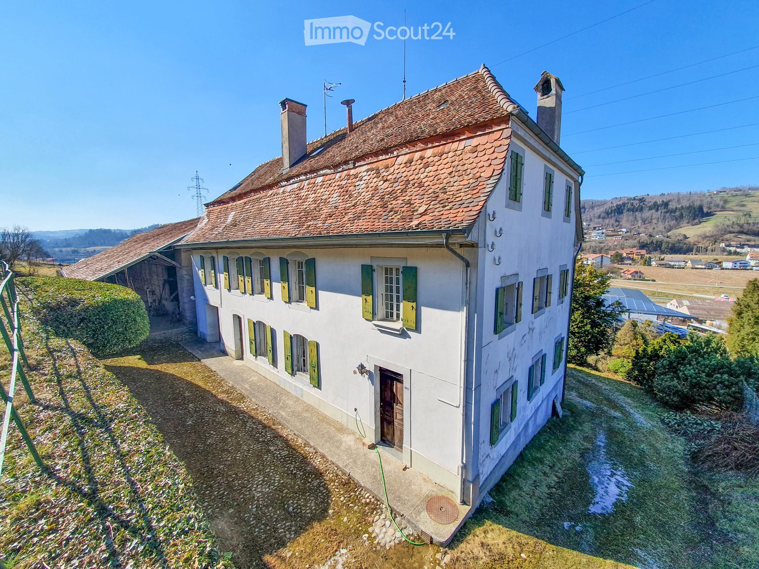 Two-story white house with green shutters, red-tiled roof, chimney, and surrounding greenery.