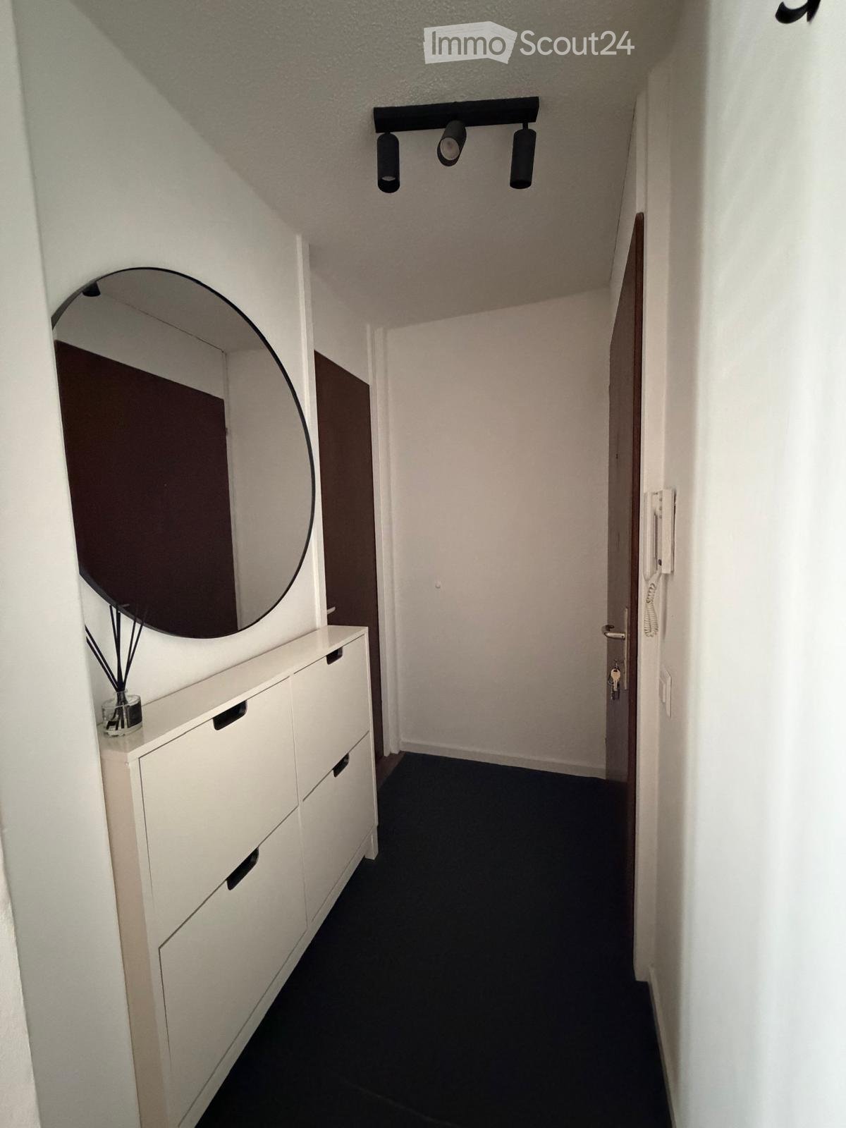 A narrow hallway with white walls, black carpet, a white cabinet with drawers, a round mirror, and a small decorative item on top of the cabinet. The hallway has a modern design with recessed lighting and a door at the end.
