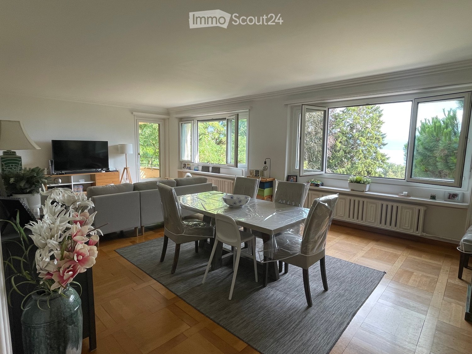 Bright living and dining area with large windows offering a scenic view, featuring a grey sectional sofa, a glass dining table with chairs, a TV on a stand, and decorative elements like a flower vase and table lamps.