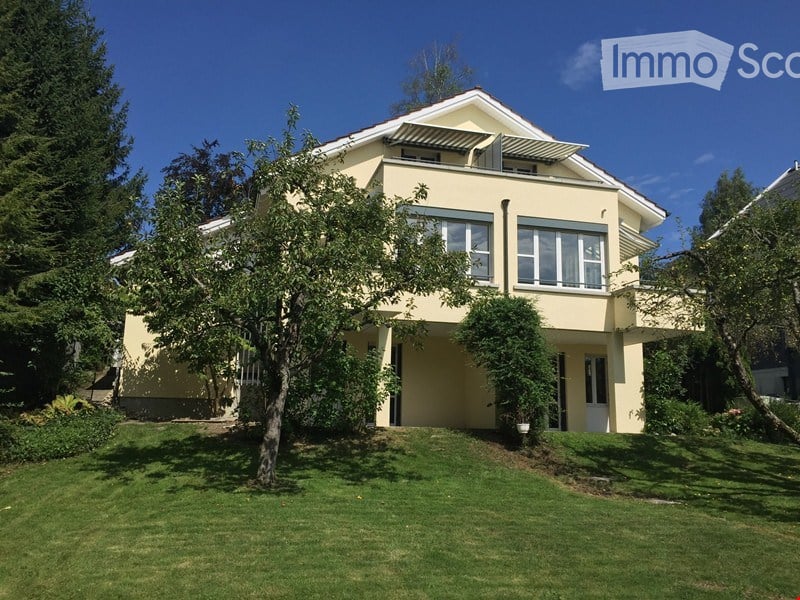 Two-story house with a light yellow exterior, large windows, and a balcony on the upper floor. The house is surrounded by a well-maintained lawn and trees.