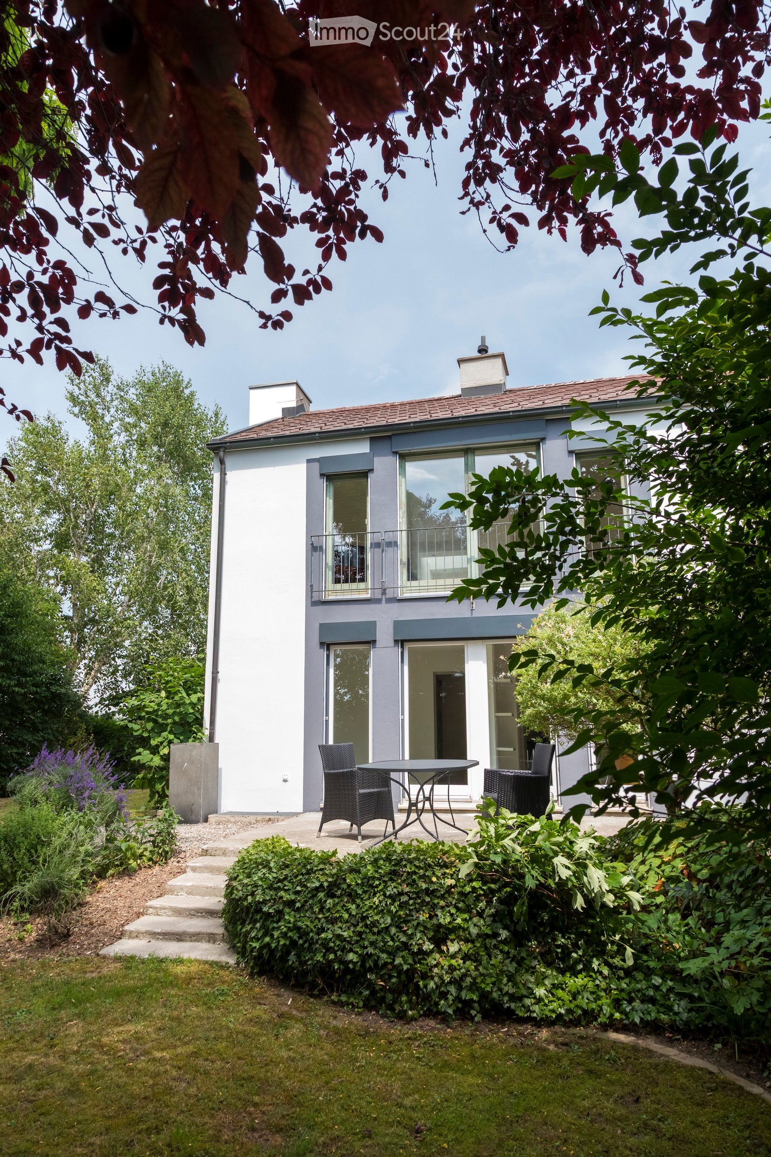 A modern two-story house with a balcony and large windows, surrounded by a well-maintained garden with a patio area featuring a table and chairs.