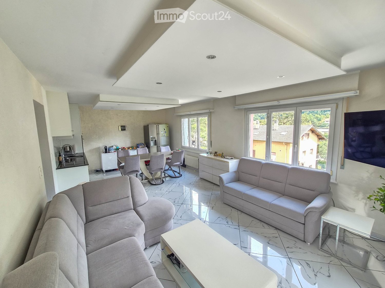 Bright living room with a grey sectional sofa, coffee table, dining area with chairs and table, kitchen area with refrigerator and cabinets, large windows with blinds, and a TV on the wall.