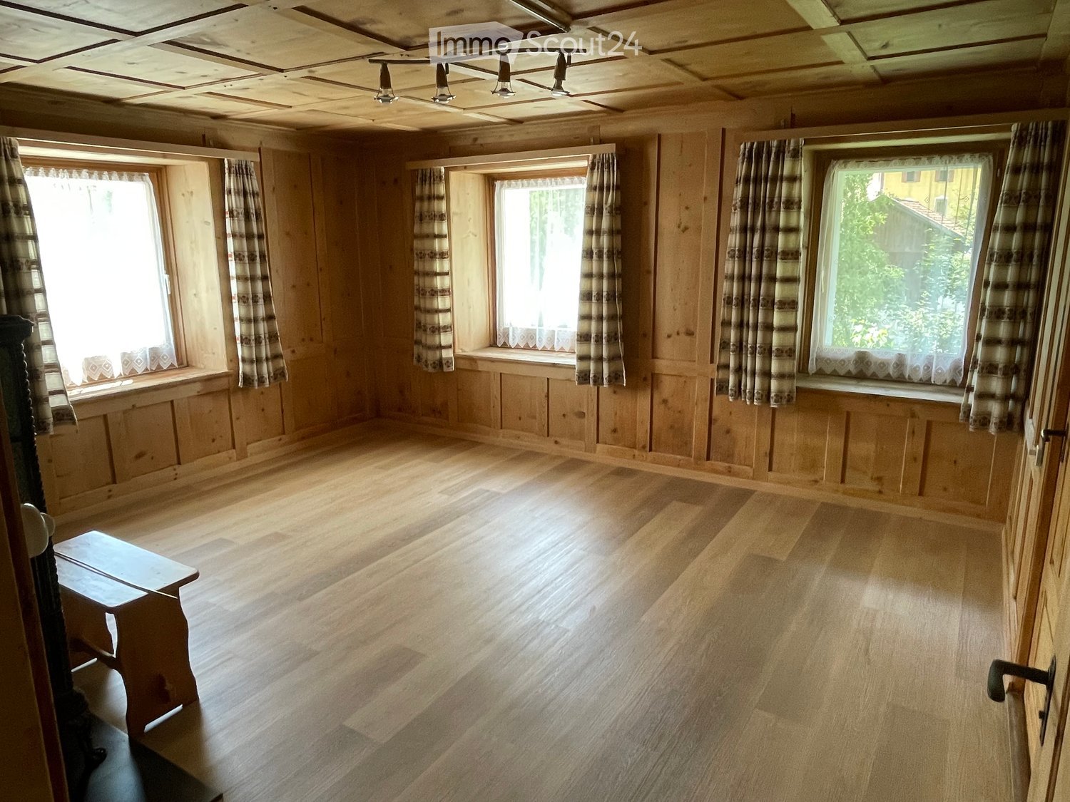 Empty room with wooden walls, wooden floor, large windows with curtains, and a small wooden bench near the door.