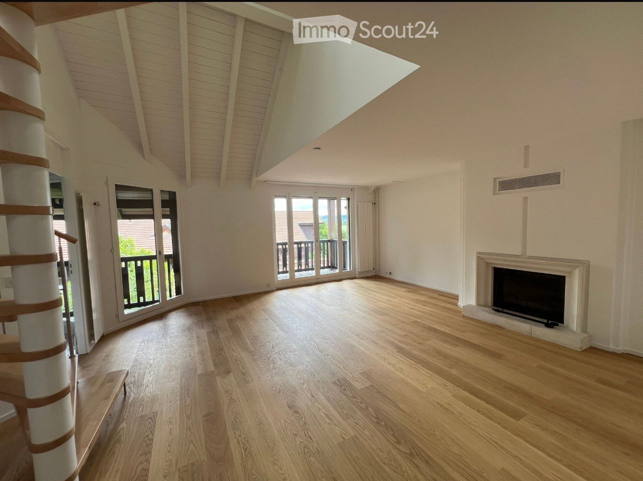 Empty living room with wooden flooring, white walls, a fireplace, and large windows leading to a balcony.