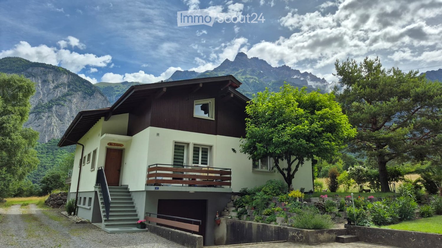 Two-story house with a balcony, garage, and surrounded by greenery and mountains.