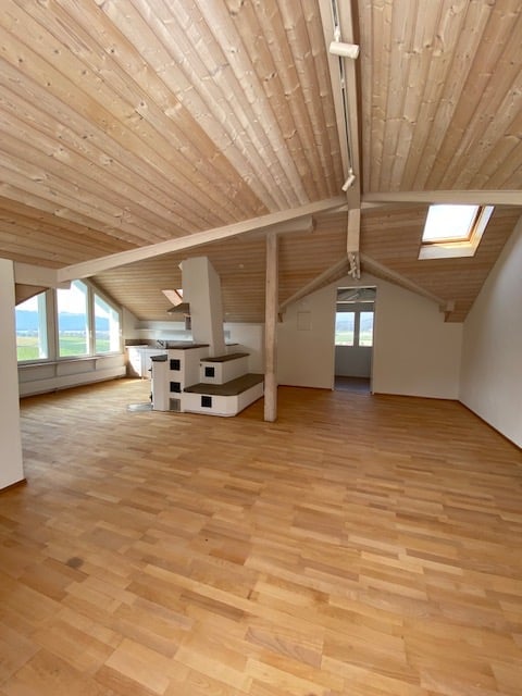 Bright, open loft-style room with wooden floors and exposed wooden beams. Features large windows, skylight, and a small staircase leading to an upper level.