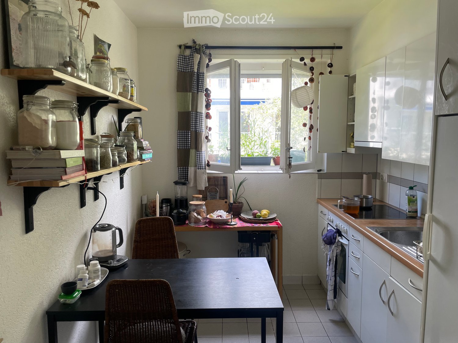 The kitchen features white cabinets, a wooden countertop, a sink, and a small dining area with a table and chairs. Shelves hold jars and books, and a window provides natural light.