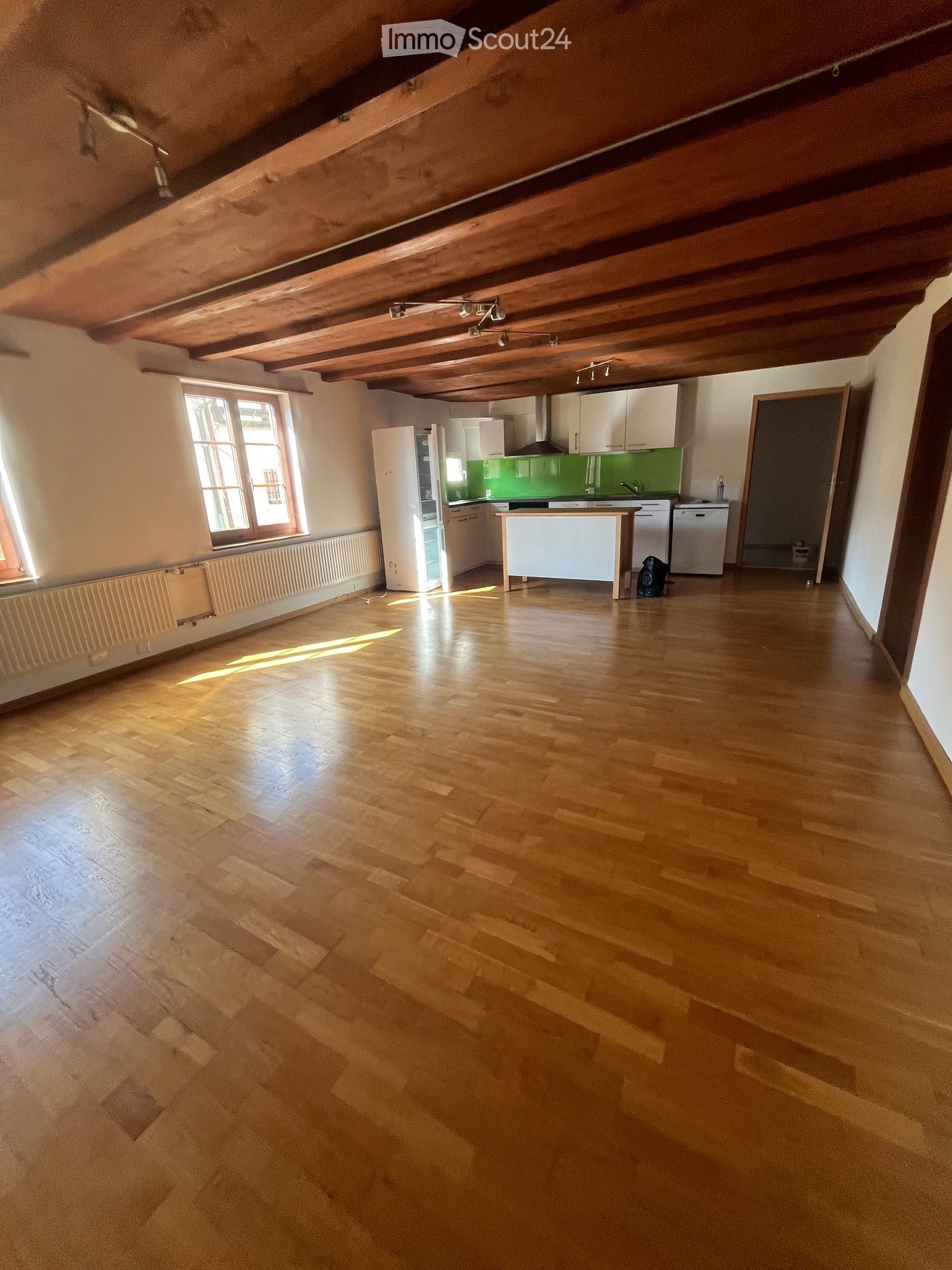 Empty room with wooden flooring, white walls, and exposed wooden ceiling beams. Features a large window with natural light and a radiator below it.