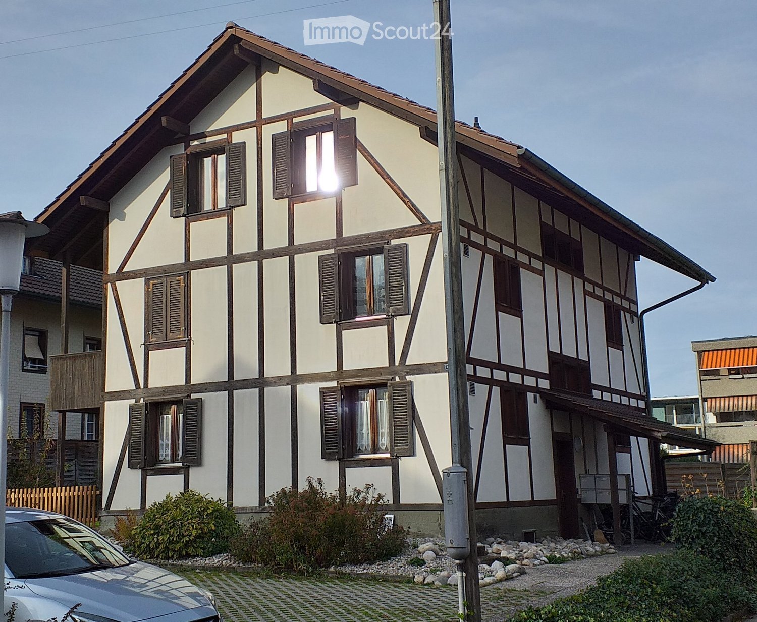 Two-story traditional house with white plaster and dark wooden beams, multiple windows with shutters, small balcony, and surrounding greenery.
