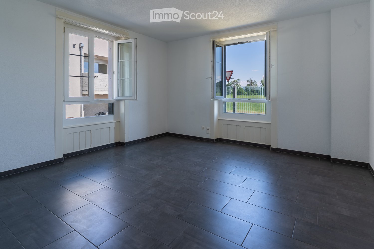 Empty room with dark tiled floor, white walls, and large windows with open shutters, showing a view of the outside area.