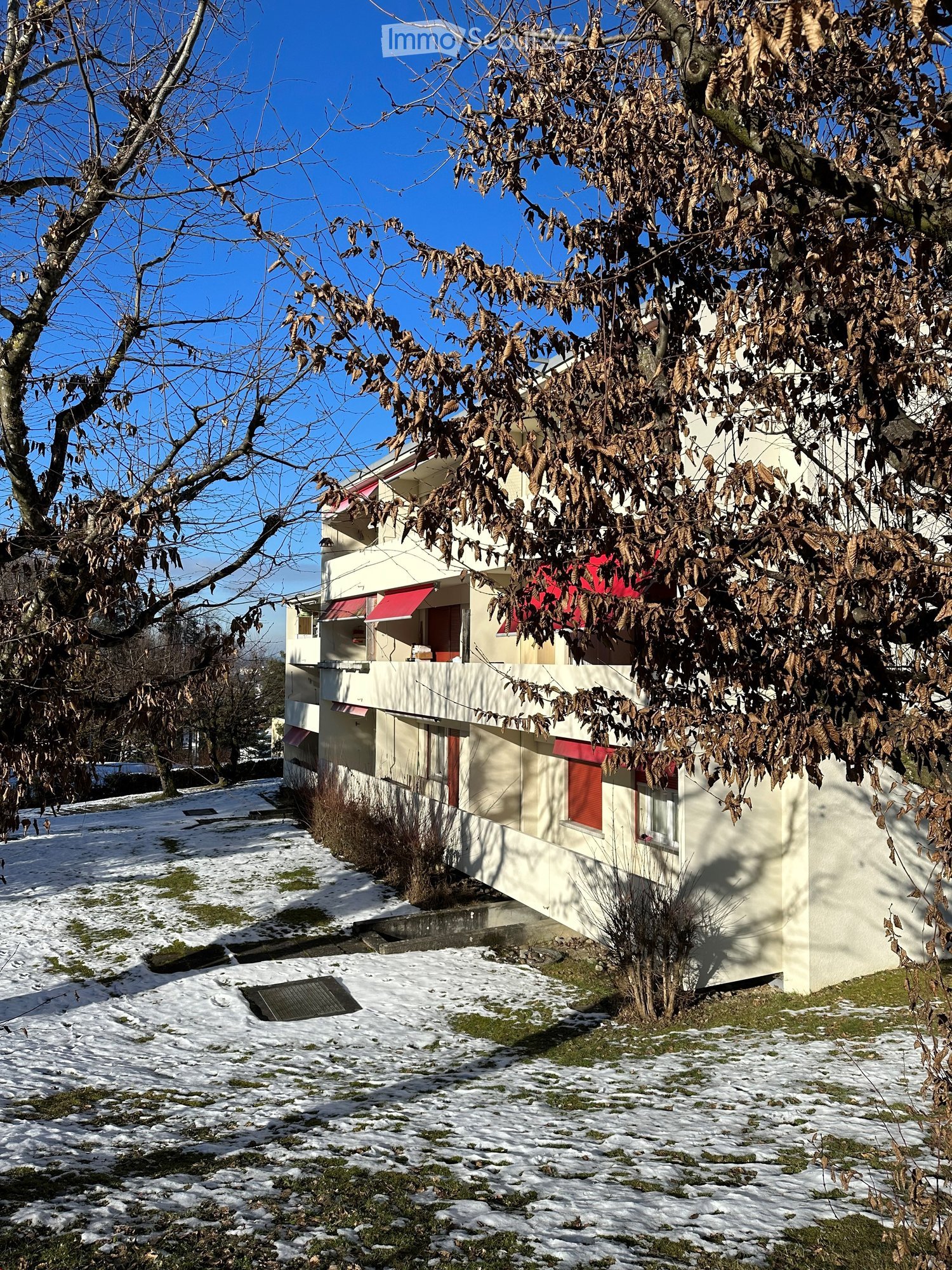Two-story house with white walls, red shutters, and a balcony partially covered by leafless trees. Snow covers the ground around the house.