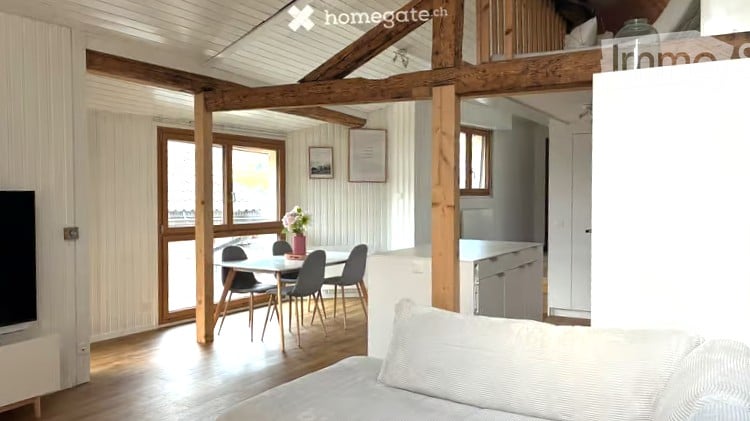 Bright, open-plan living and dining area with exposed wooden beams, large windows, a sofa, dining table with chairs, and a small kitchen area with cabinets and a refrigerator.
