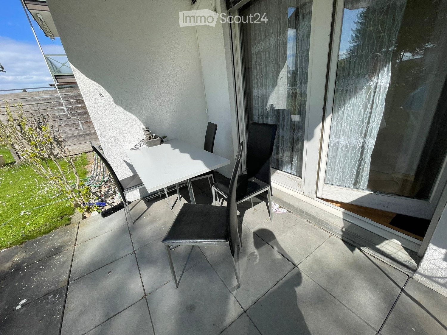 Balcony with a white table and four black chairs, overlooking a grassy area with a wooden deck and a small tree.