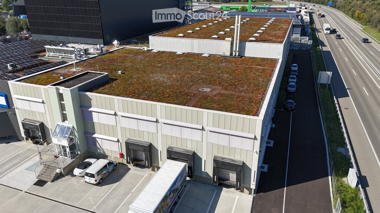 Modern industrial building with a green roof, multiple loading docks, and a parking area with several vehicles.
