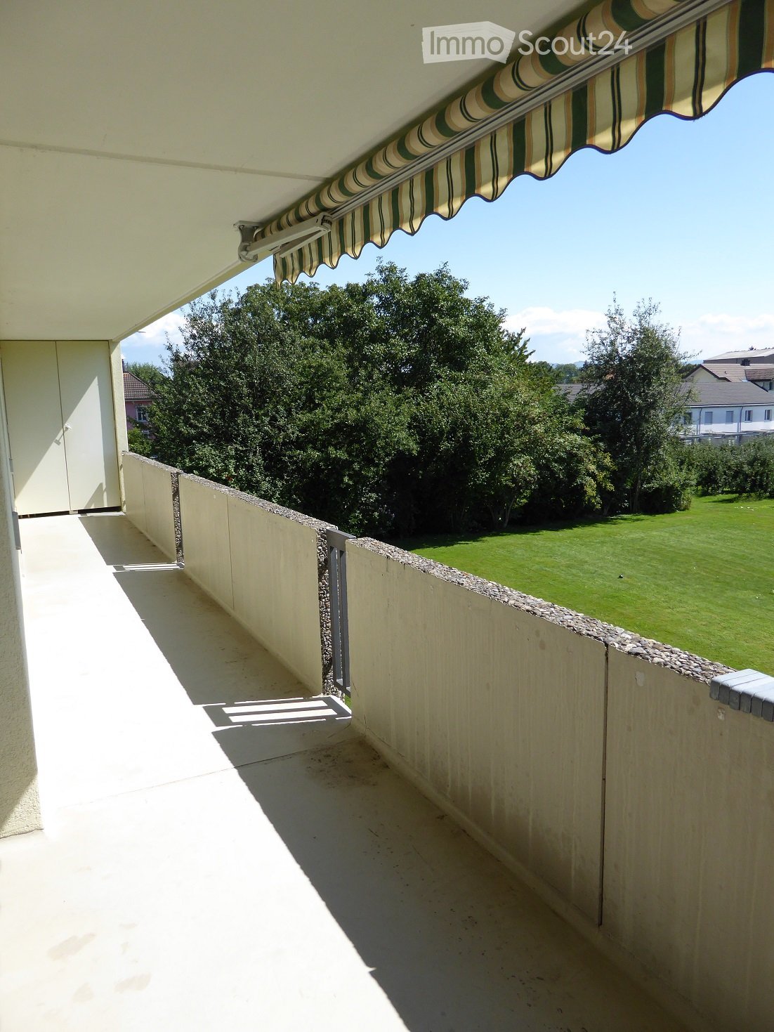 Balcony with a striped awning, low stone wall, and view of a green lawn and trees.