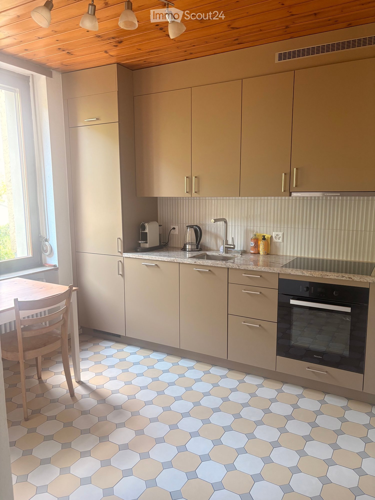 Modern kitchen with light-colored cabinets, a hexagonal tile floor, a small dining area with a table and chair, and various appliances including a coffee maker, oven, and sink.