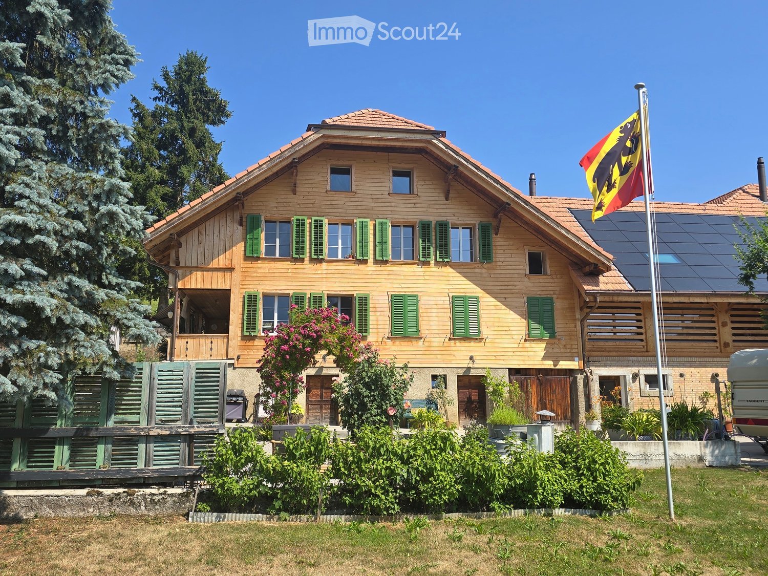 Two-story wooden house with green shutters, multiple windows, and a flagpole with a flag. The house has a well-maintained garden with various plants and trees.