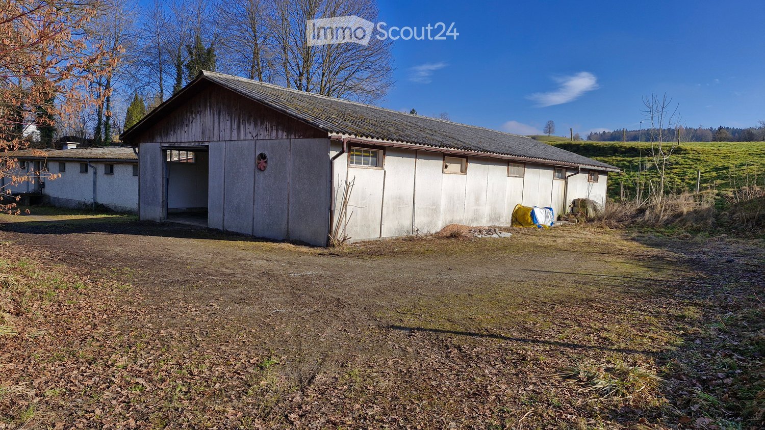 A rustic, single-story building with a barn-like structure, featuring a large open doorway, small windows, and a metal roof. The surrounding area includes a dirt ground and some scattered items near the building.