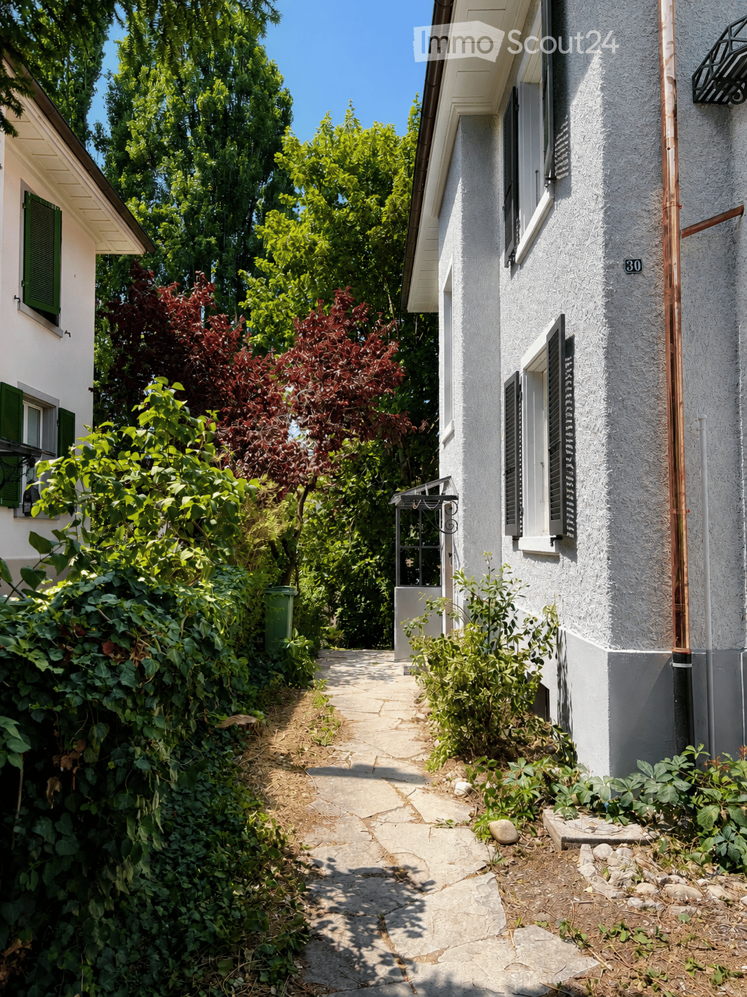 A narrow garden pathway lined with greenery leads to a white house with green shutters and a small balcony. The house has a textured facade and a visible address number30.