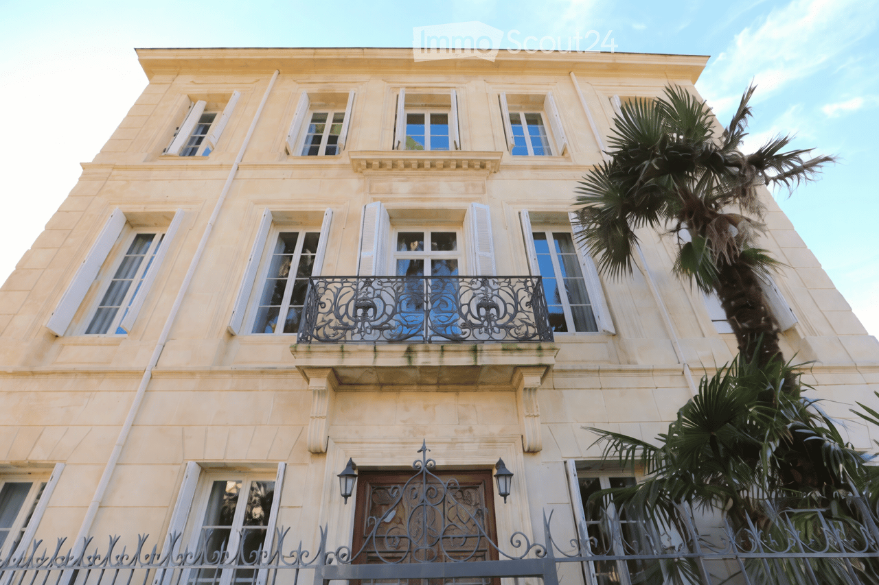 A multi-story building with light-colored stone facade, multiple windows with shutters, ornate wrought-iron balcony, and palm trees in front.