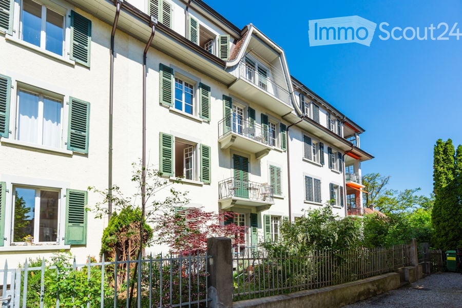Multi-story residential building with white facade, green shutters, multiple balconies, and surrounding greenery.