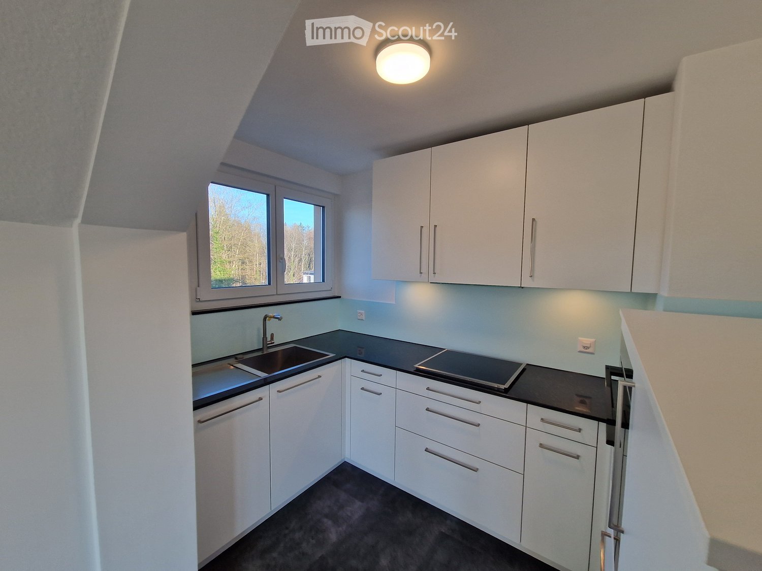 Modern kitchen with white cabinets, black countertops, a sink, and a stove, featuring a large window with a view of trees.