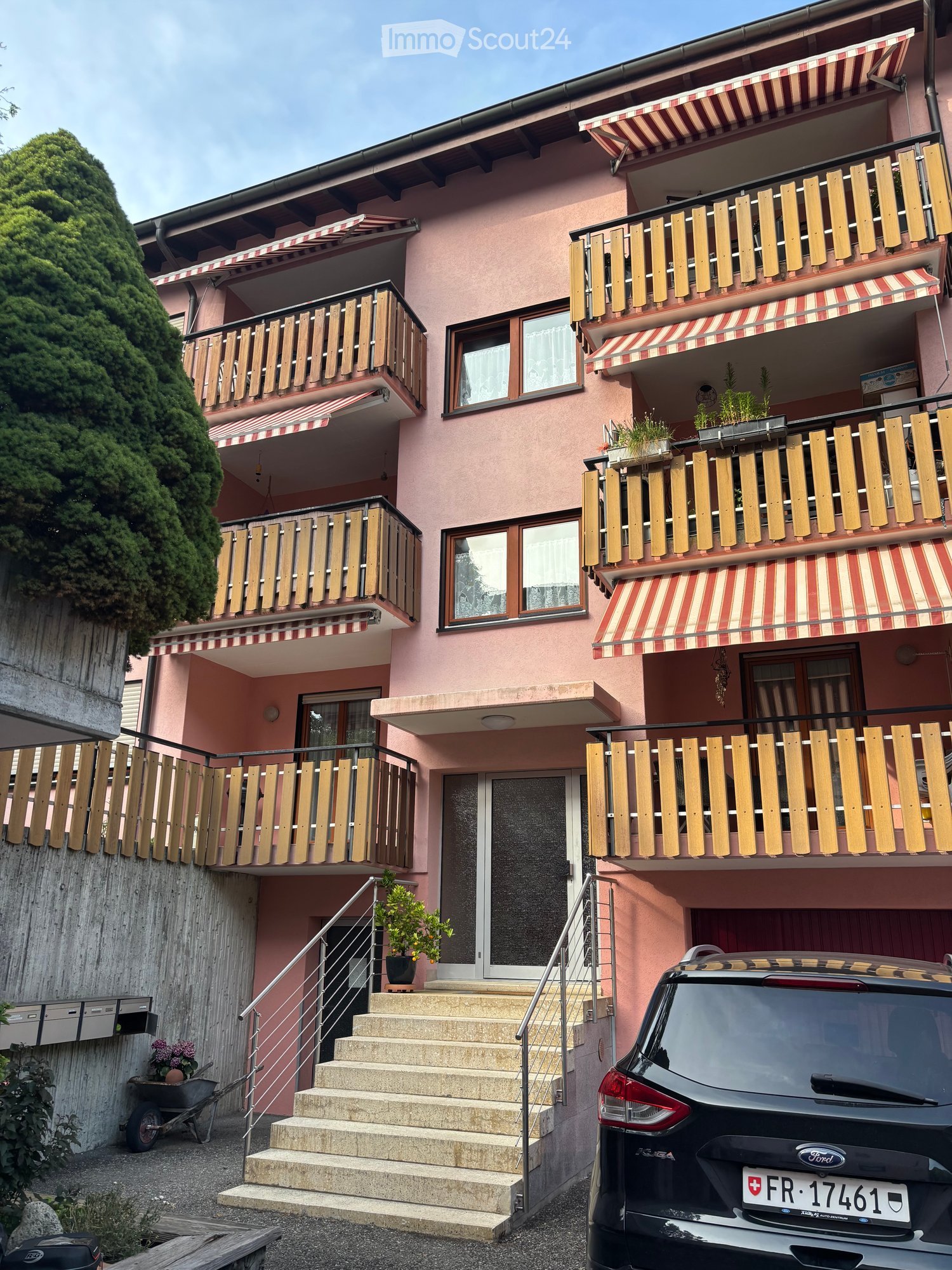 Pink multi-story residential building with multiple balconies, striped awnings, and a staircase leading to the entrance. A car is parked in front.