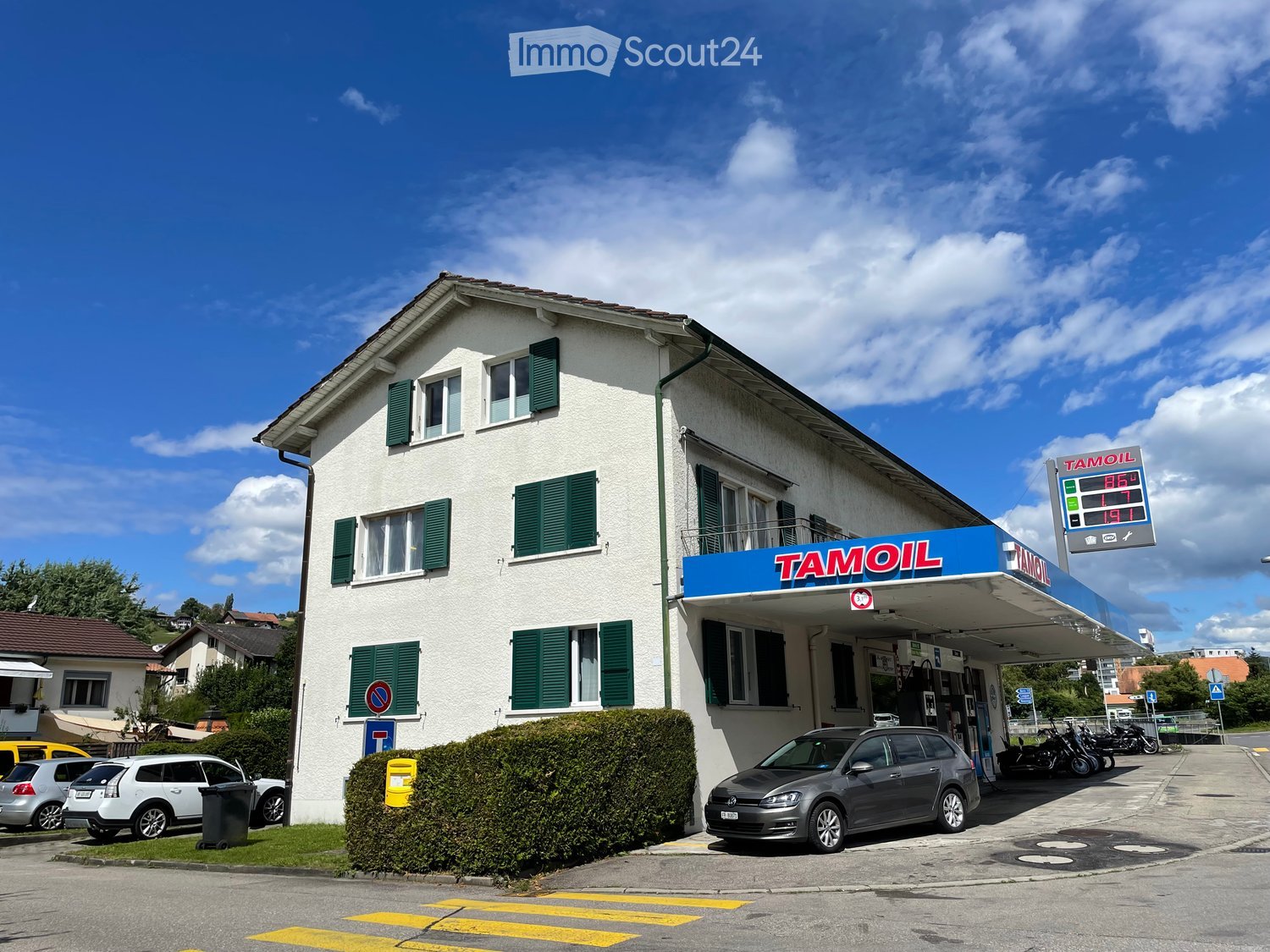 Two-story white building with green shutters, Tamoil gas station attached, several parked cars, and a clear blue sky.