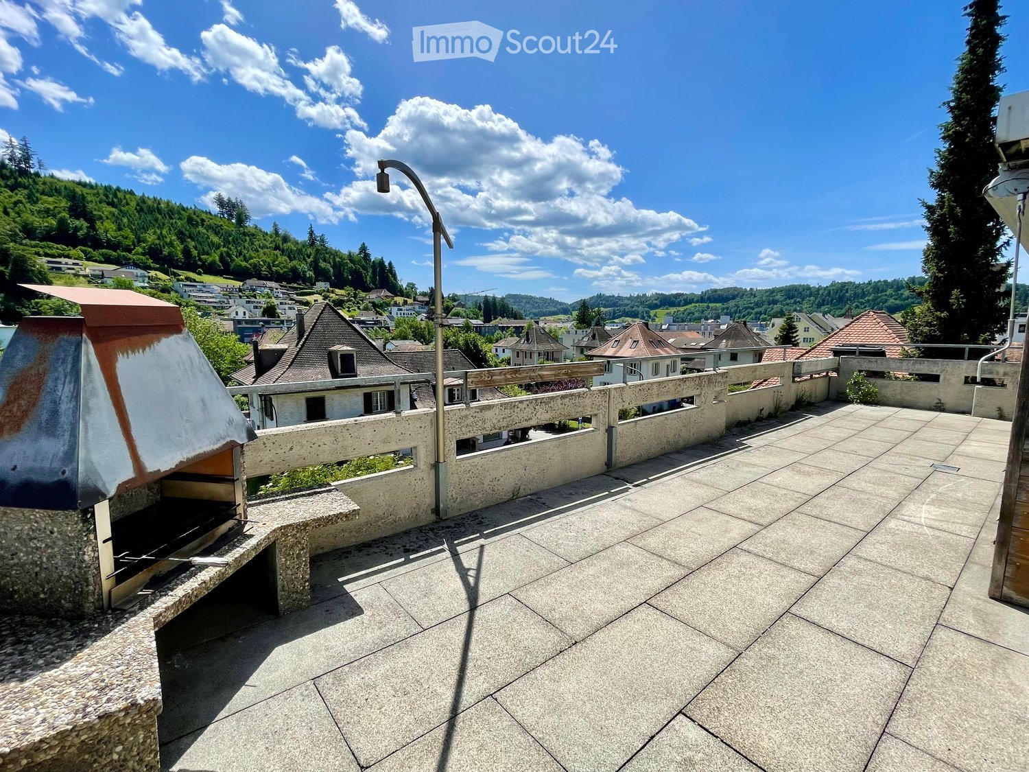 Roof terrace with concrete tiles, low stone walls, and a view of a town and surrounding hills.