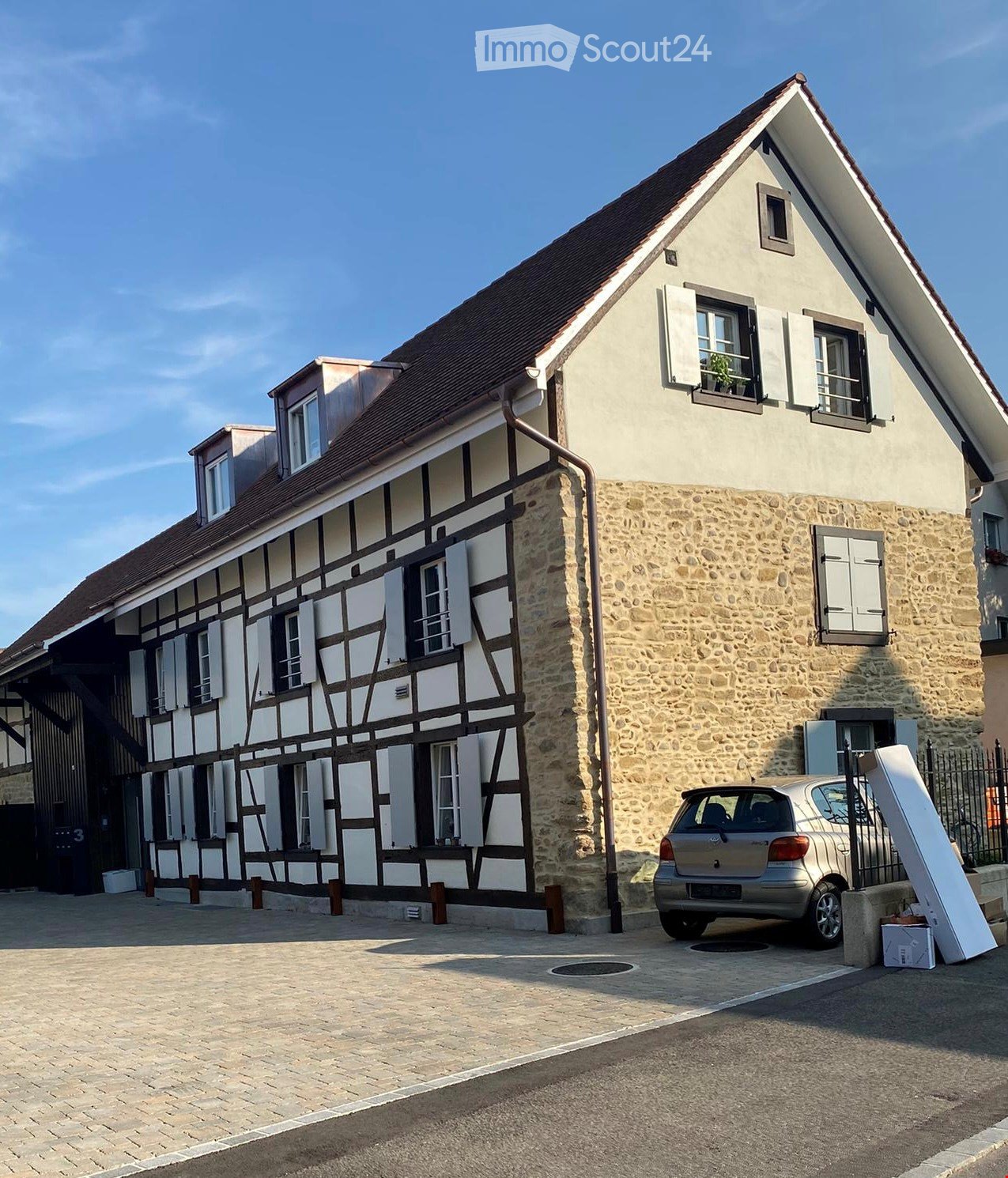 Two-story house with a combination of stone and timber framing, multiple windows with shutters, and a parked car in front.