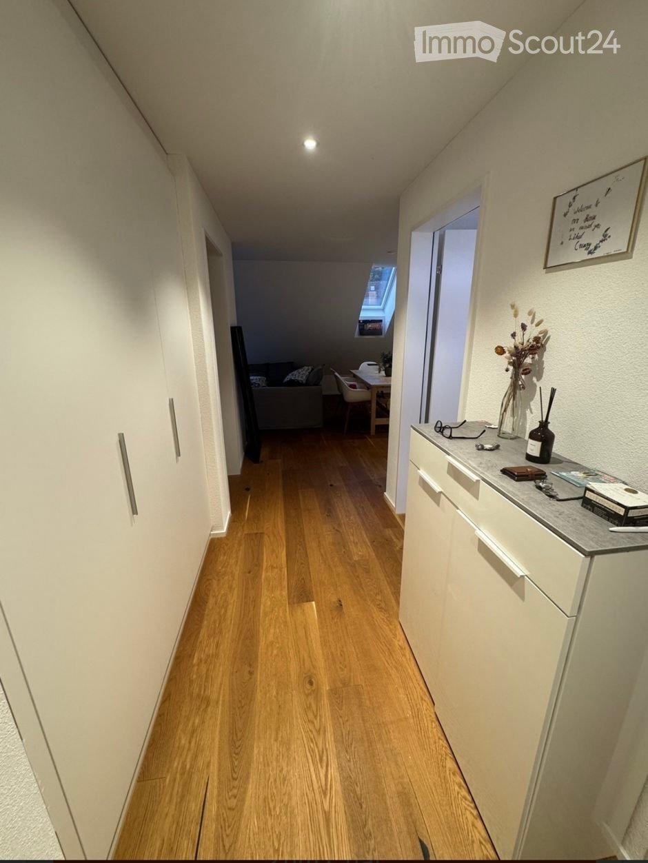 A narrow hallway with wooden flooring, white walls, and built-in storage cabinets. A small table with a vase and decorative items is visible on the right side, and a glimpse of a living area with a sofa and dining table can be seen at the end of the hallw