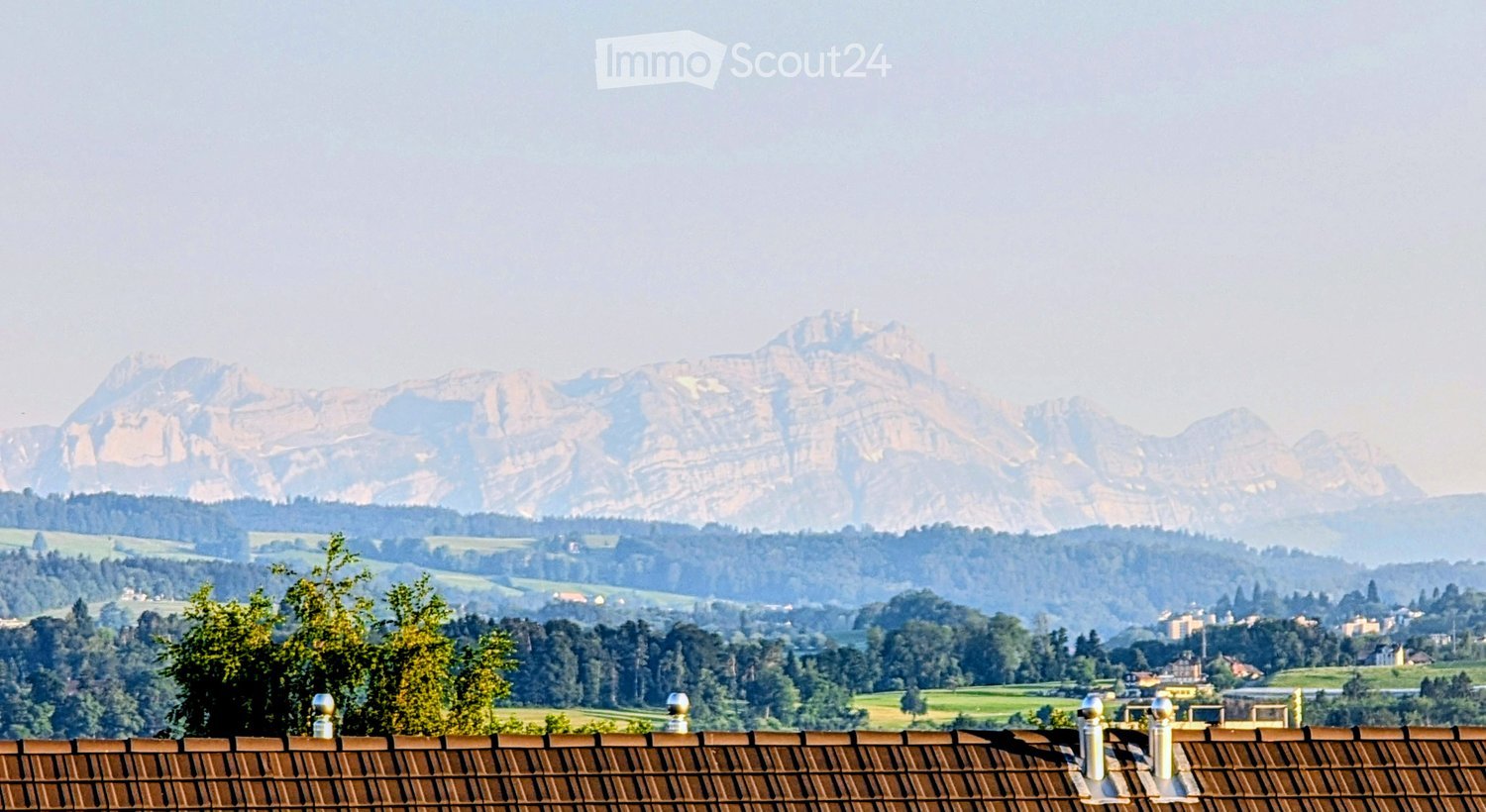 View from a rooftop terrace showing a panoramic landscape with rolling hills, dense forests, and distant mountains under a hazy sky.