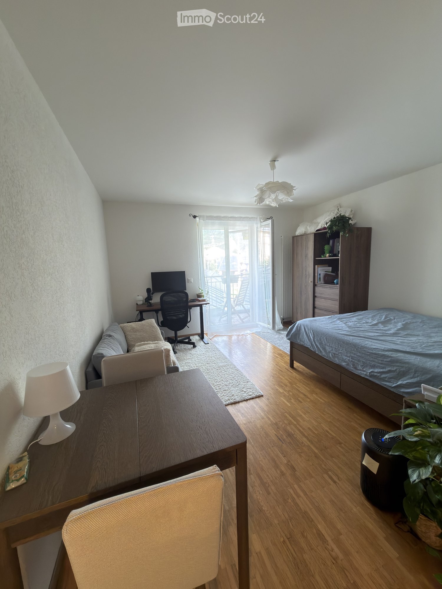 A well-lit bedroom with a bed, desk, and sitting area. Features include a wooden floor, white walls, a large window with a view, and modern furniture.