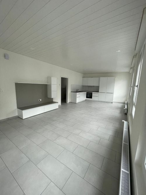 Modern, minimalist kitchen and living area with white cabinets, a built-in bench, and grey tiled flooring.
