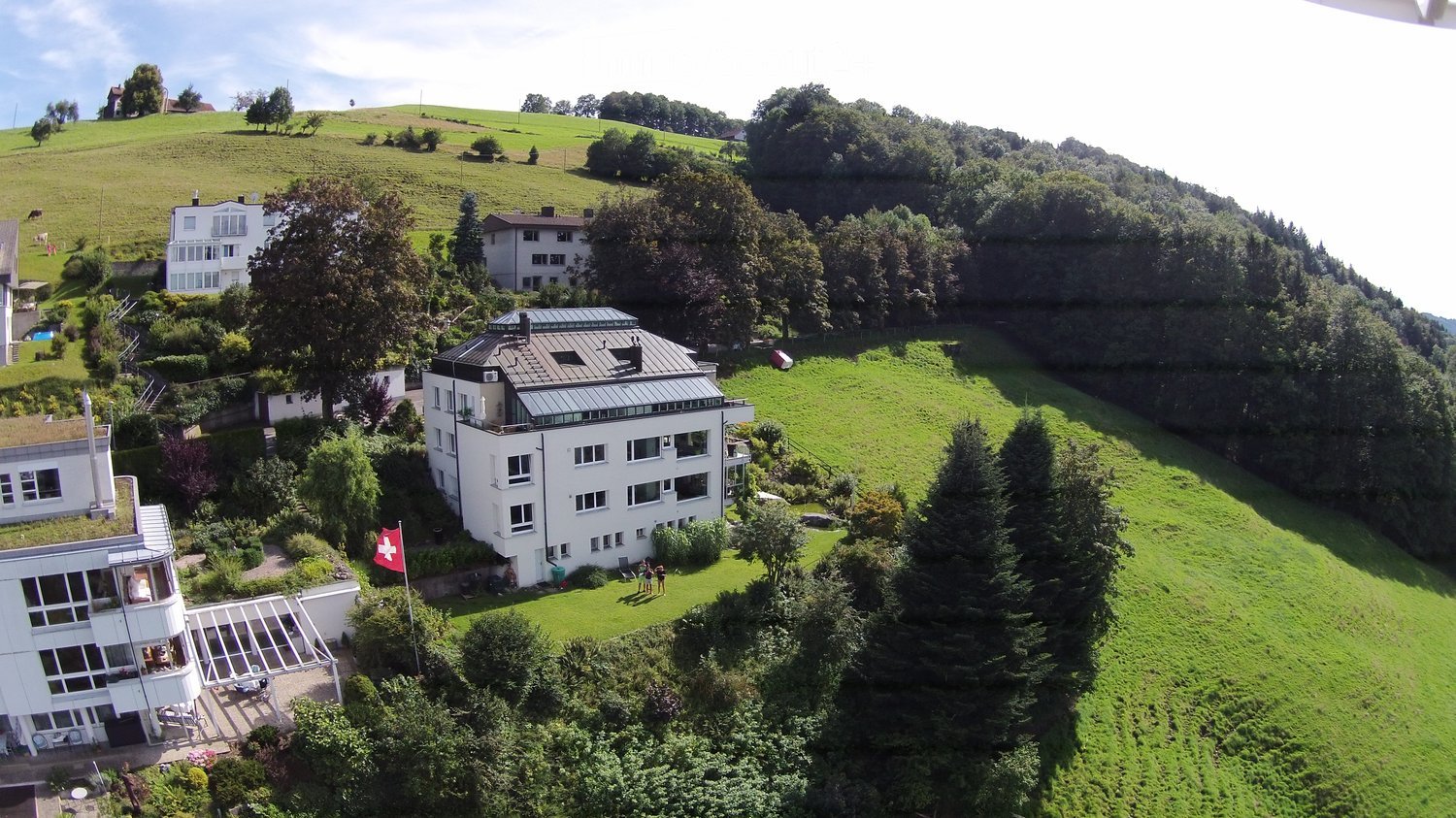 Aerial view of a modern white building with a flat roof and large windows, surrounded by lush green hills and trees. The building has a Swiss flag displayed in front.