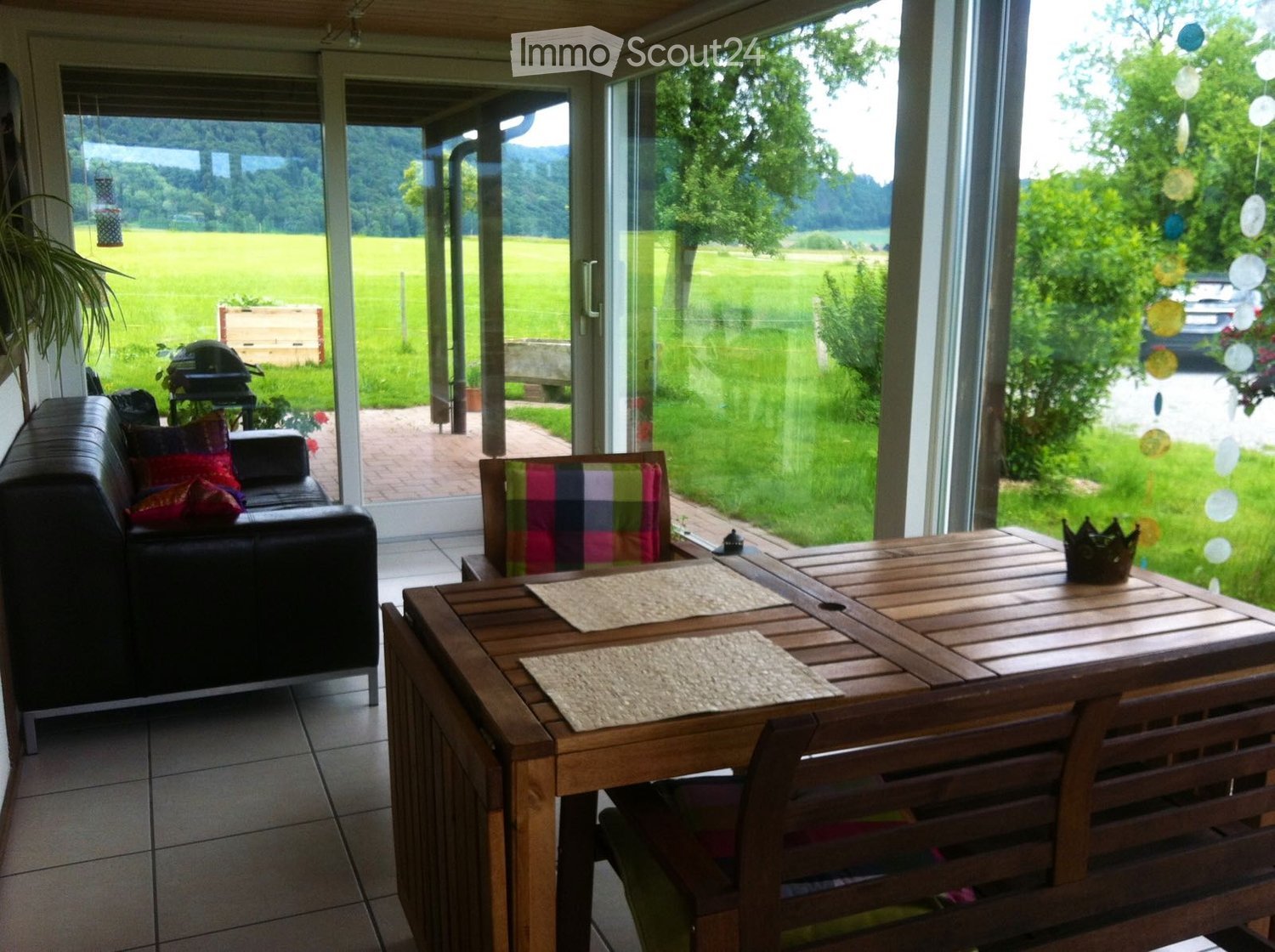 Bright living room with large glass doors opening to a green outdoor area, featuring a black leather sofa with colorful cushions, a wooden dining table with placemats and a decorative crown centerpiece, and a colorful striped chair.