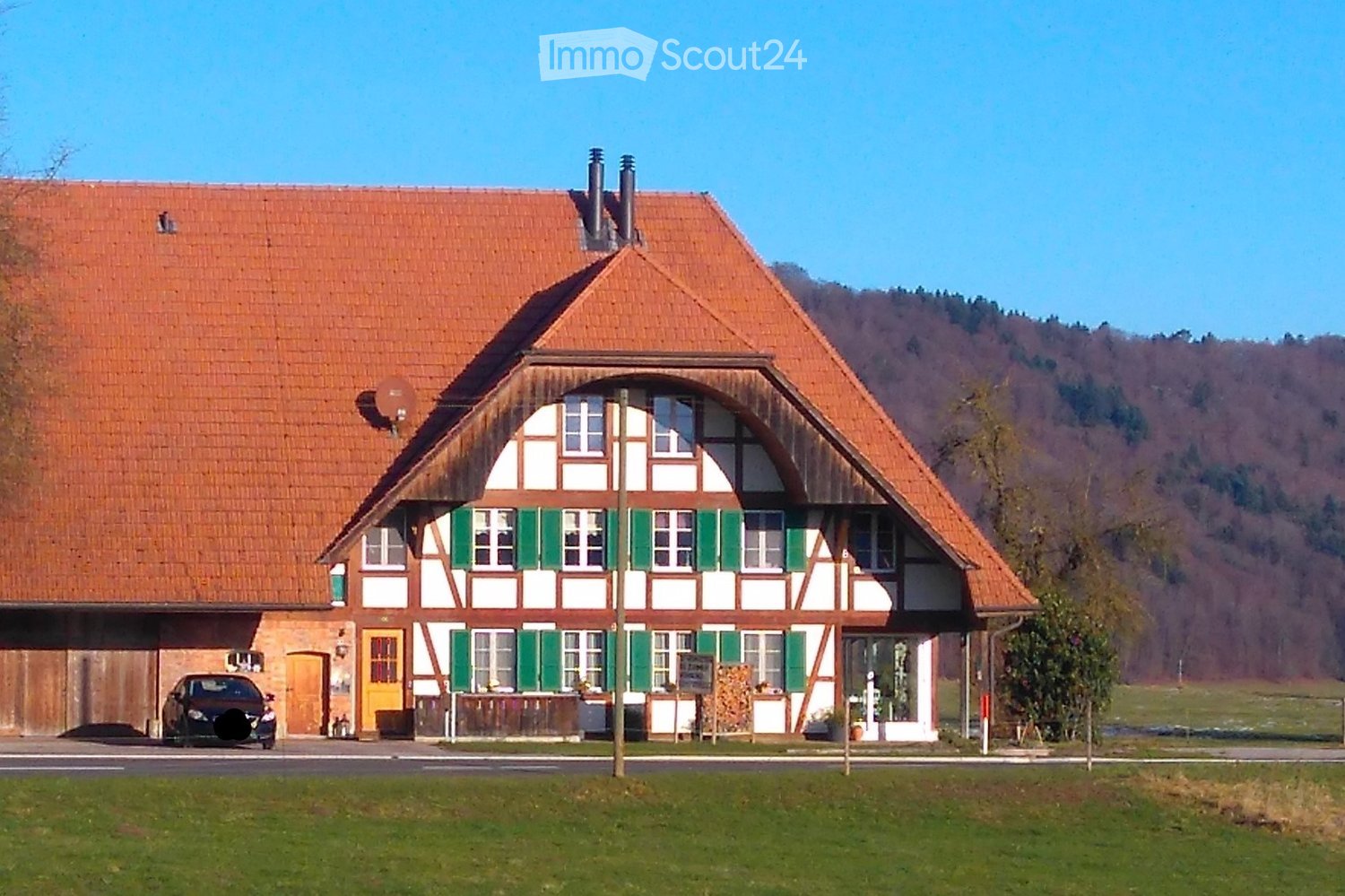 Traditional two-story house with a red-tiled roof, white and green timber framing, multiple windows, and a garage. A car is parked in front, and the house is surrounded by a grassy lawn.