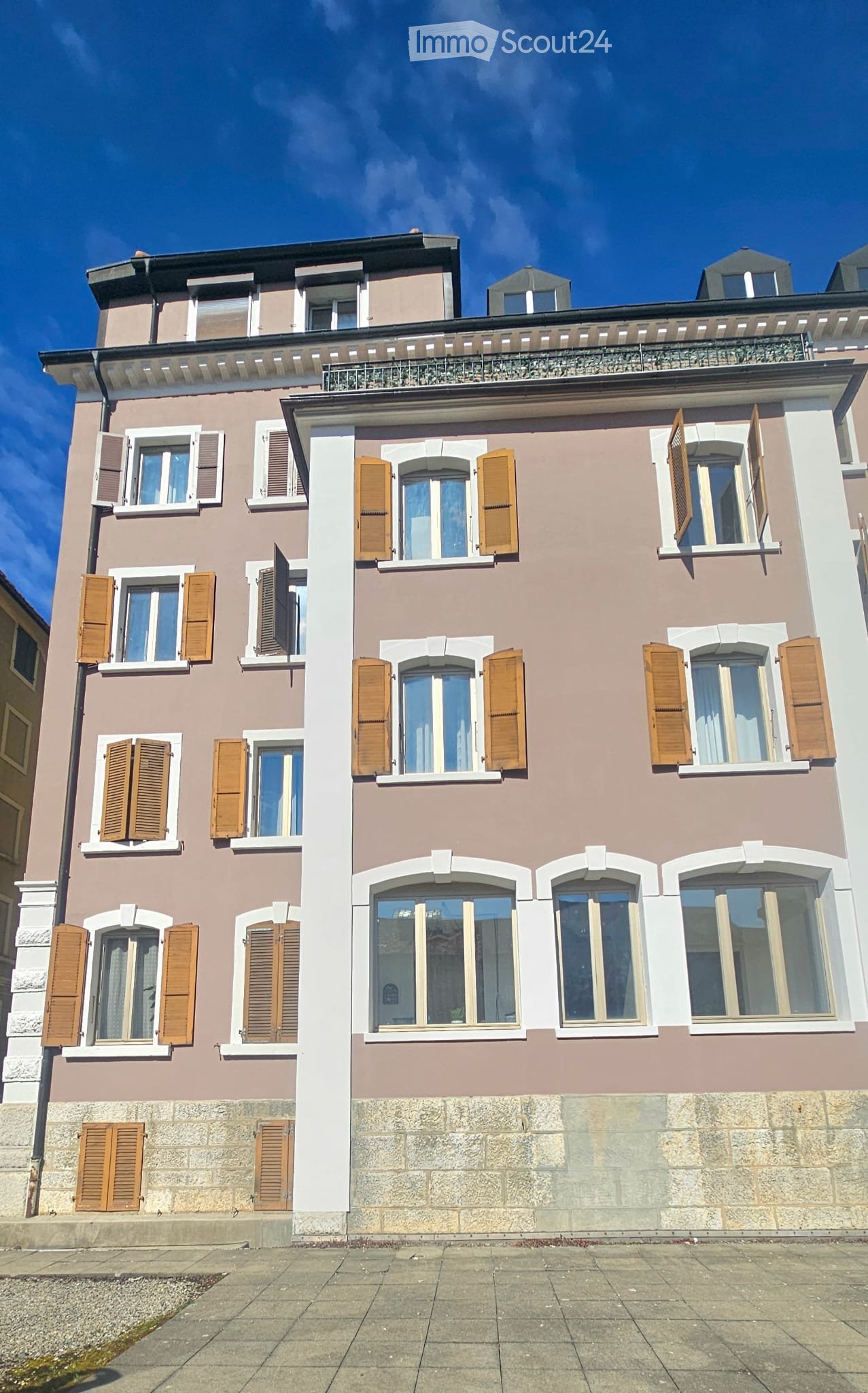 A three-story residential building with beige and brown facade, multiple windows with wooden shutters, and a balcony on the top floor.