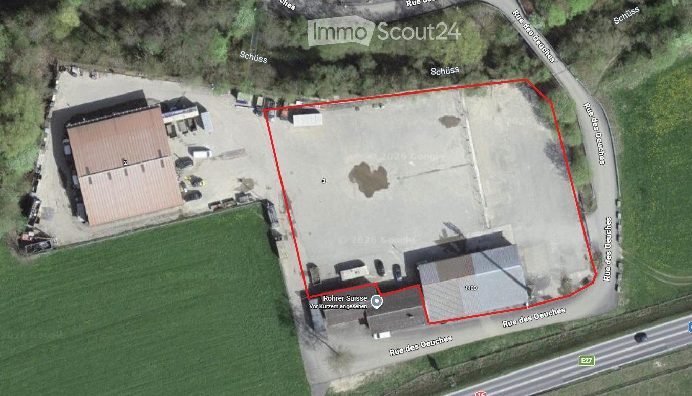 Aerial view of a building complex with a large paved area, surrounded by roads and greenery. The complex includes multiple structures and parking spaces.
