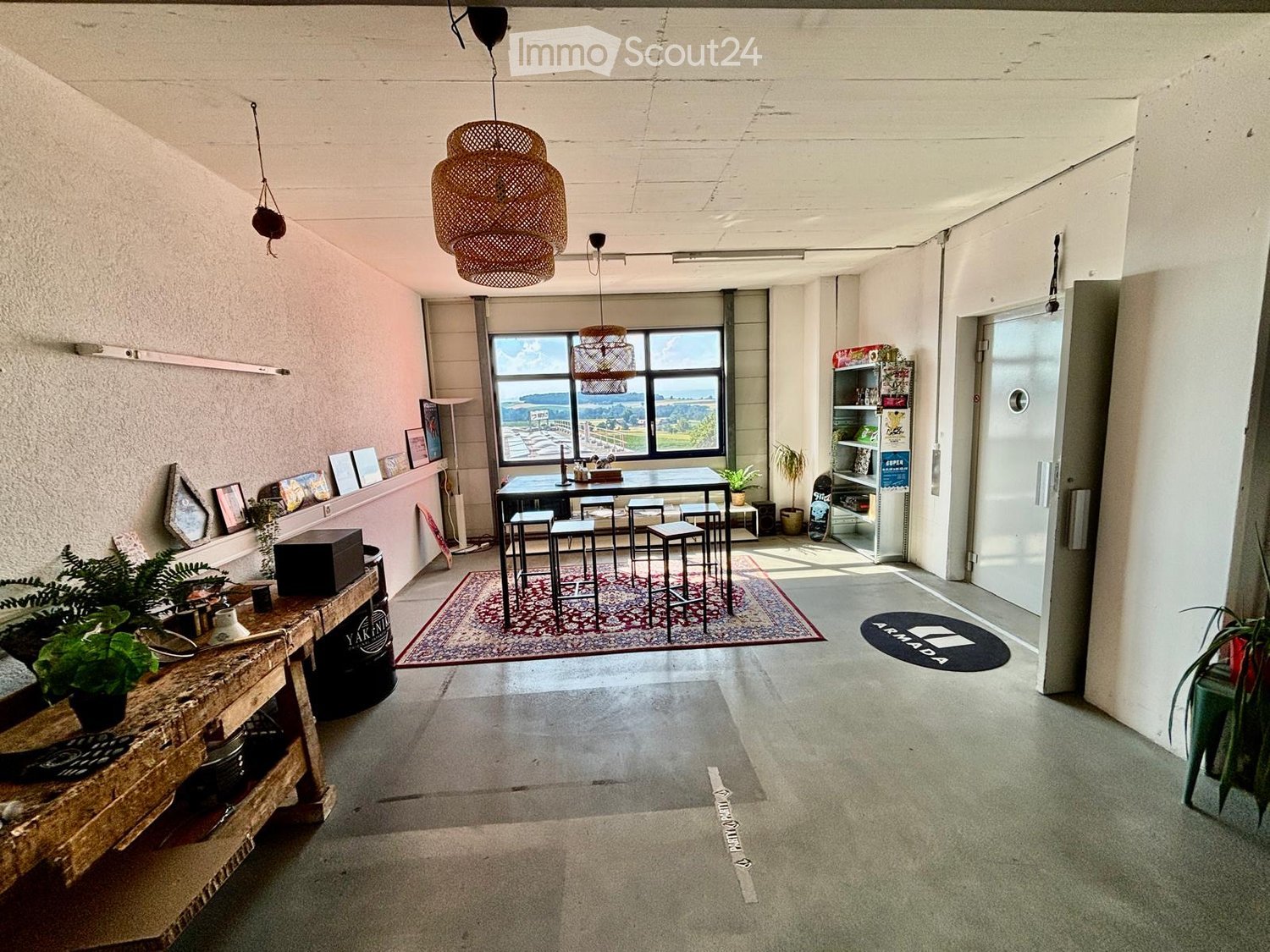 Open-plan space with a dining area, bar counter, and shelves. Features a large window with a scenic view, wicker pendant lights, and potted plants.