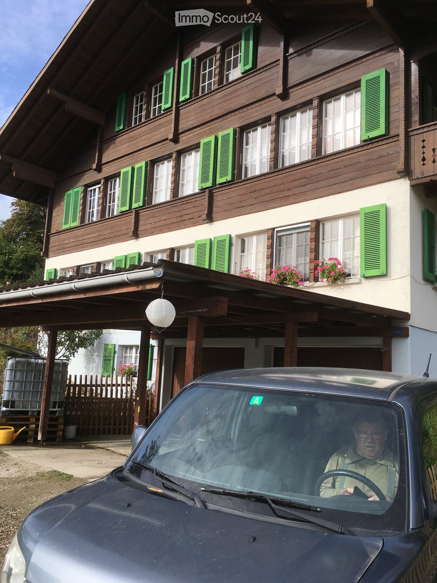 Two-story wooden house with green shutters, white-framed windows, and a covered entrance area. The house features a balcony with flower boxes and a parked car in the foreground.